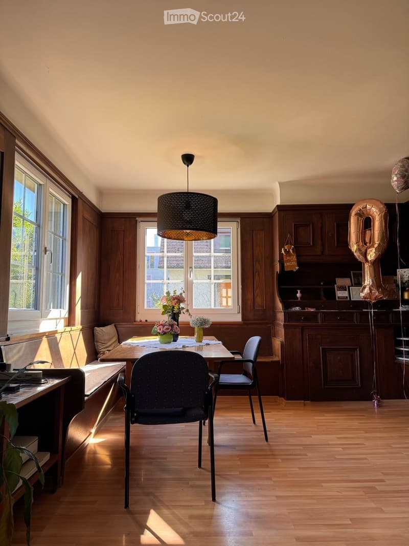 Brightly lit dining area with wooden floors, large windows, a round wooden table with chairs, a pendant light fixture, and a cabinet with decorative items and balloons.