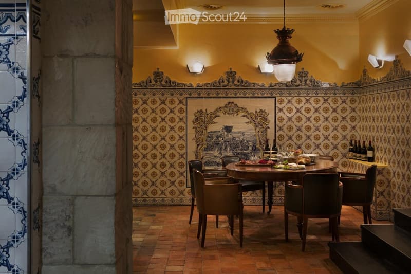 Elegant dining area with a round wooden table surrounded by upholstered chairs, set with wine bottles, plates, and glasses. The walls feature intricate decorative tiles and a chandelier hangs from the ceiling.