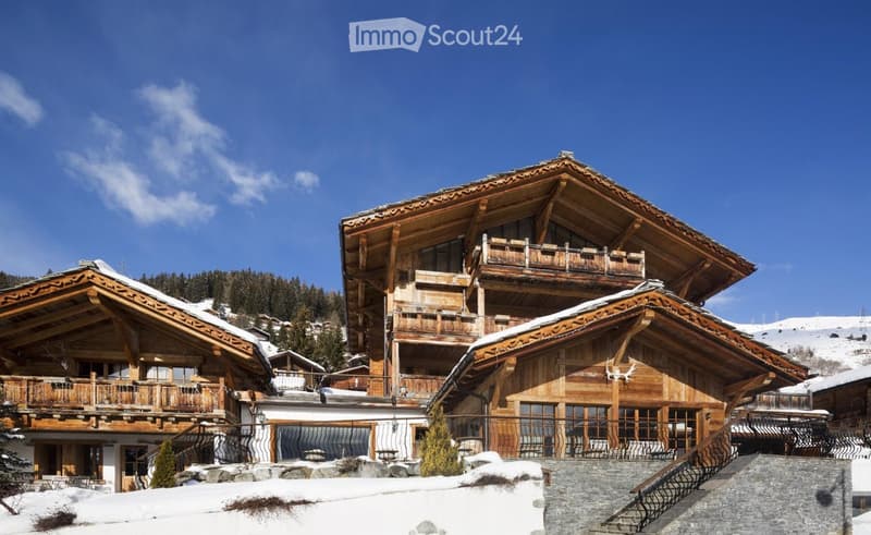 A large, multi-story wooden chalet with snow-covered roofs and balconies, surrounded by snow and trees.