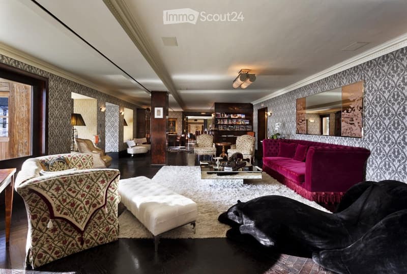 Elegant living room with patterned wallpaper, dark hardwood floors, and a mix of plush seating including a red velvet sofa, floral-patterned armchairs, and a white ottoman. A coffee table sits in the center, flanked by additional seating and a large mirro