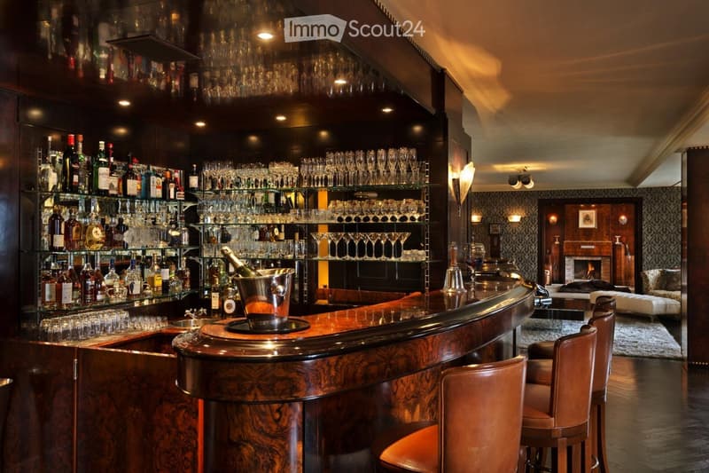 Elegant bar area with a curved wooden counter, leather bar stools, wine rack filled with bottles and glasses, ice bucket, and a cozy seating area with a fireplace and sofa.