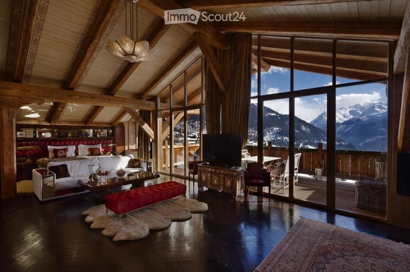 Luxurious bedroom with exposed wooden beams, large windows offering mountain views, a red upholstered bed, a white sofa, a coffee table, a red bench, a television, and a chandelier.