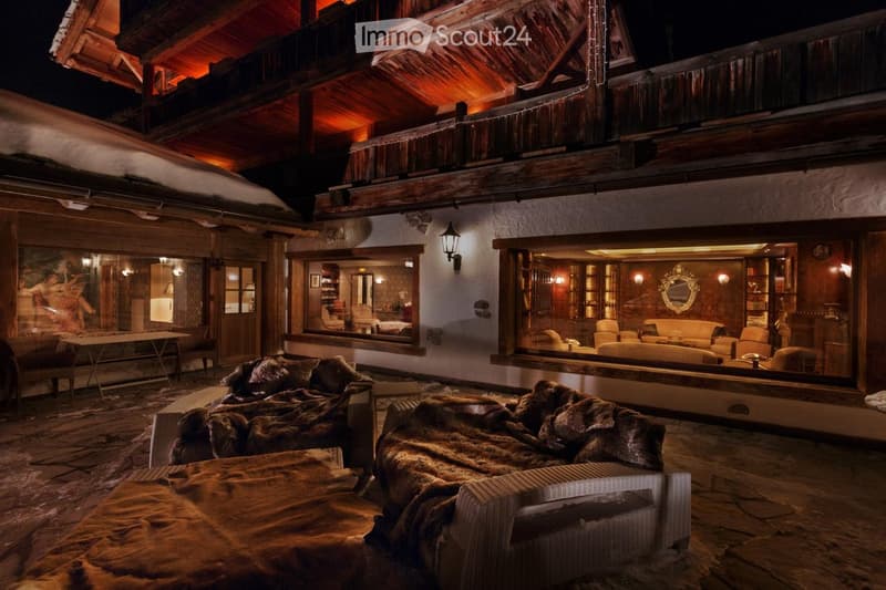 An outdoor lounge area with stone flooring, wooden beams, and fur-covered seating, connected to an indoor living space with large windows and warm lighting.