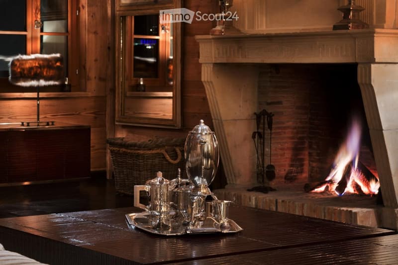 Cozy living room with a lit fireplace, wooden walls, a dark wooden table with silver teapot and cups, fireplace tools, and a wicker basket nearby.