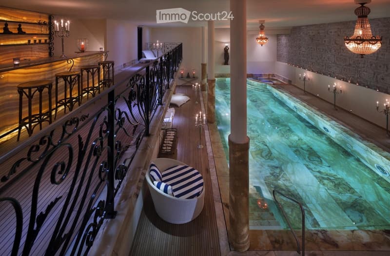 Luxurious indoor pool area with marble floors, ornate columns, chandeliers, and decorative seating, featuring a long swimming pool with a bar area on the upper level.