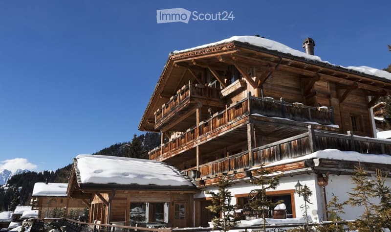 A large, multi-story wooden chalet with snow-covered roofs and balconies, surrounded by snow-covered trees and mountains.