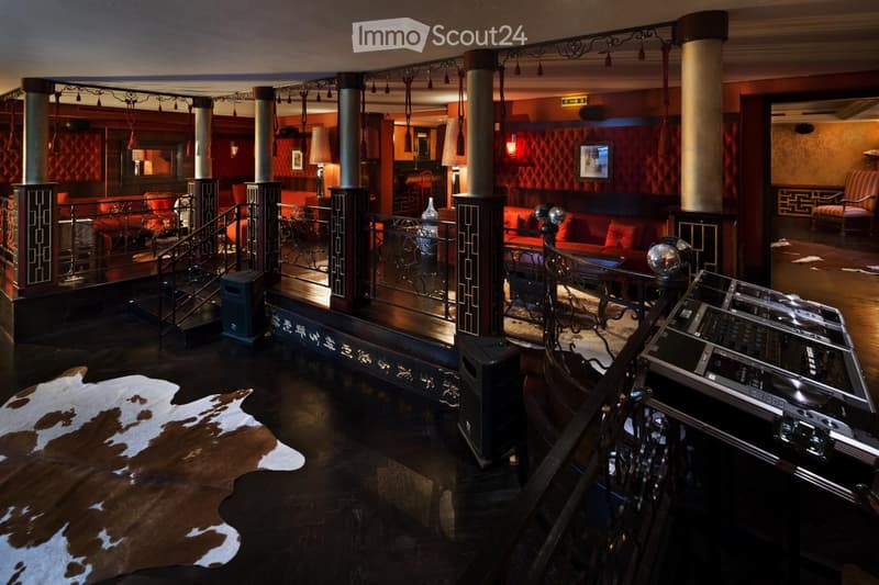 Elegant lounge area with dark wooden floors, red velvet walls, ornate columns, and a staircase leading to an upper level. Features include a cowhide rug, lamps, a DJ booth, and seating areas with sofas and chairs.