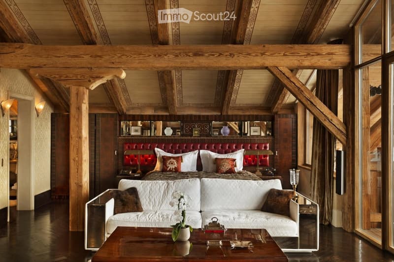 Luxurious bedroom with exposed wooden beams, a large red leather headboard bed, a white sofa, a coffee table with a flower vase, and wall-mounted lamps.