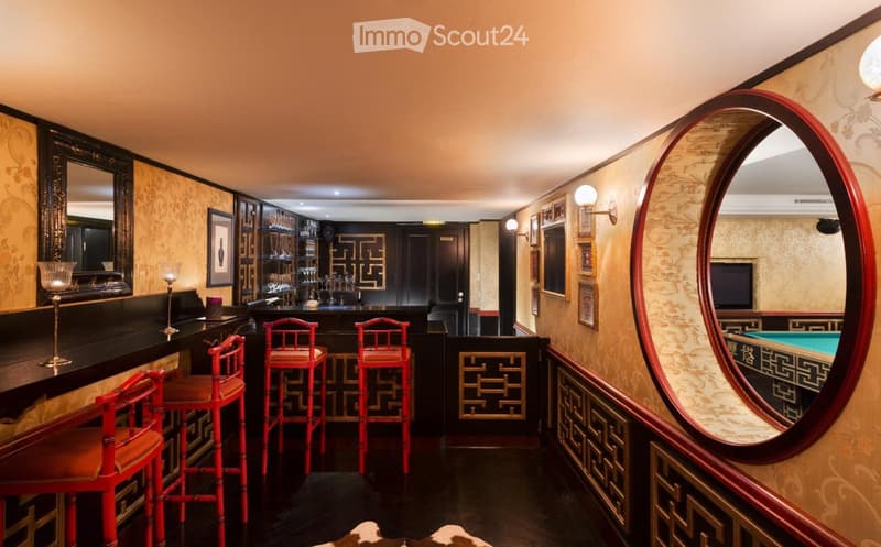 Elegant bar area with red stools, a large round mirror, decorative wall art, and a television. The space features ornate wallpaper and a dark polished floor.