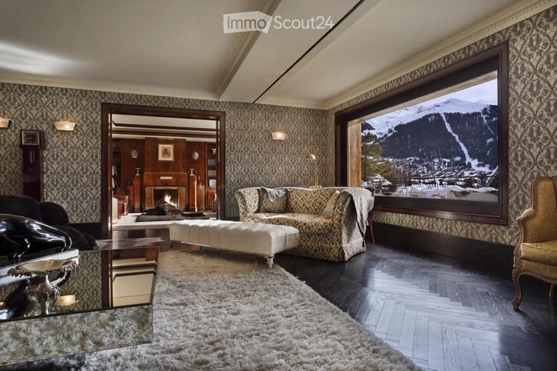 Elegant living room with patterned wallpaper, large window showcasing a snowy mountain view, a patterned sofa, ottoman, armchair, coffee table, fireplace, and a mirror.