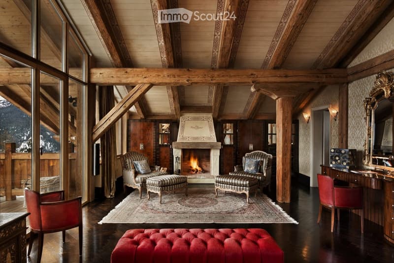 Luxurious living room with exposed wooden beams, a stone fireplace, patterned armchairs, a red velvet ottoman, and a large mirror. The room features a wooden desk and a patterned rug.