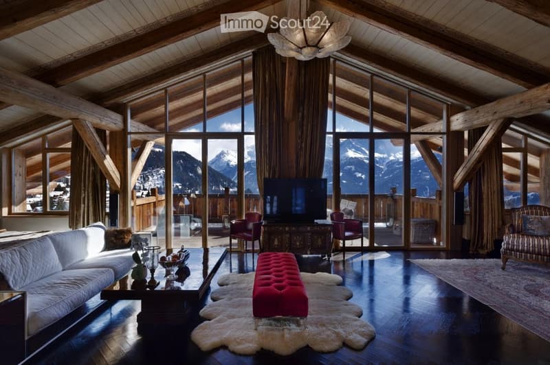 A spacious living room with exposed wooden beams, large windows offering a mountain view, a white sofa, a red bench, a coffee table, and a television set.