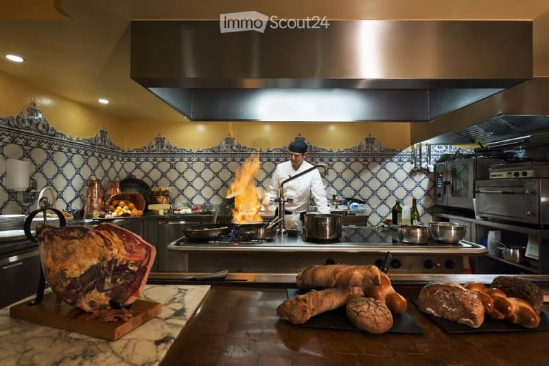 A chef is cooking in a professional kitchen with a large stove, pots, pans, and a variety of food items including bread and cured meat. The kitchen features a decorative tile backsplash and a large exhaust hood.