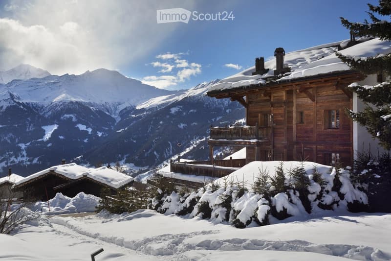 A wooden chalet with snow-covered roof and surrounding snow-covered trees, set against a backdrop of snow-capped mountains.