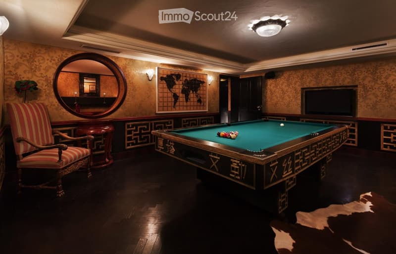 Elegant room with dark wood flooring, a large pool table, a striped armchair, a round wooden table, a large round mirror, and a television mounted on the wall.