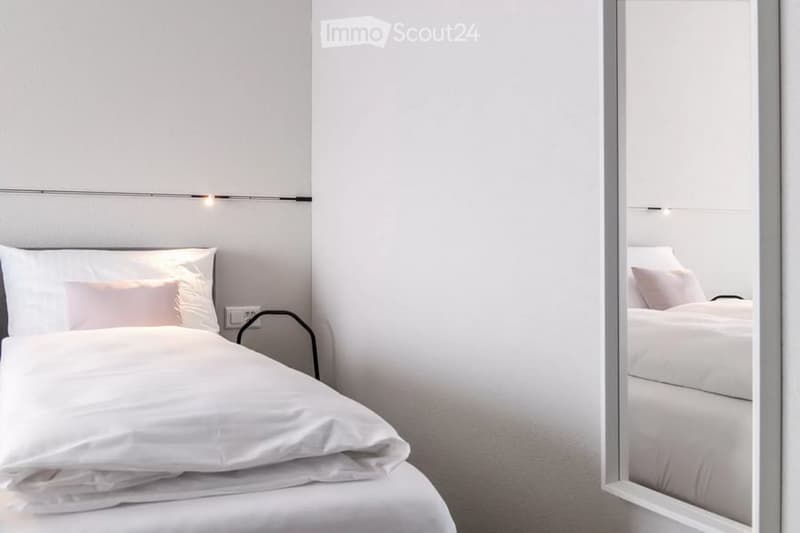 A modern, minimalist bedroom with a neatly made bed, white bedding, and a large mirror reflecting the bed.