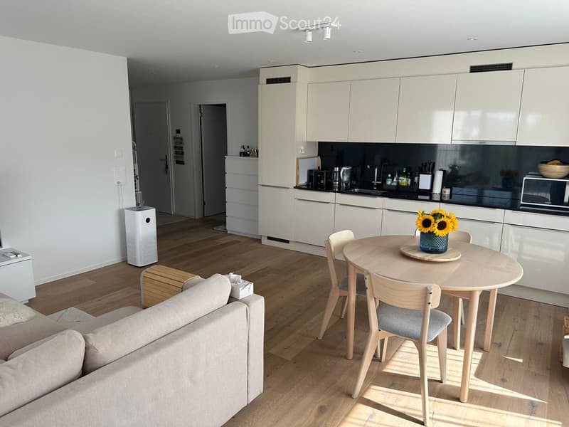 Open-plan living and dining area with light wood flooring, white cabinets, a sofa, a round dining table with chairs, and a vase with sunflowers on the table.