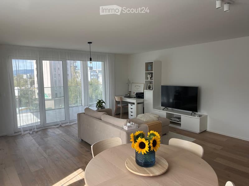 Modern living room with large windows and balcony access, featuring a sofa, coffee table, dining table with chairs, TV, desk, and decorative plant.