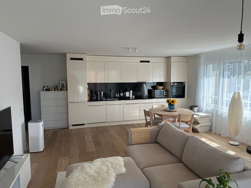 Modern living and dining area with a grey sofa, coffee table, dining table with chairs, white kitchen cabinets, and various appliances.