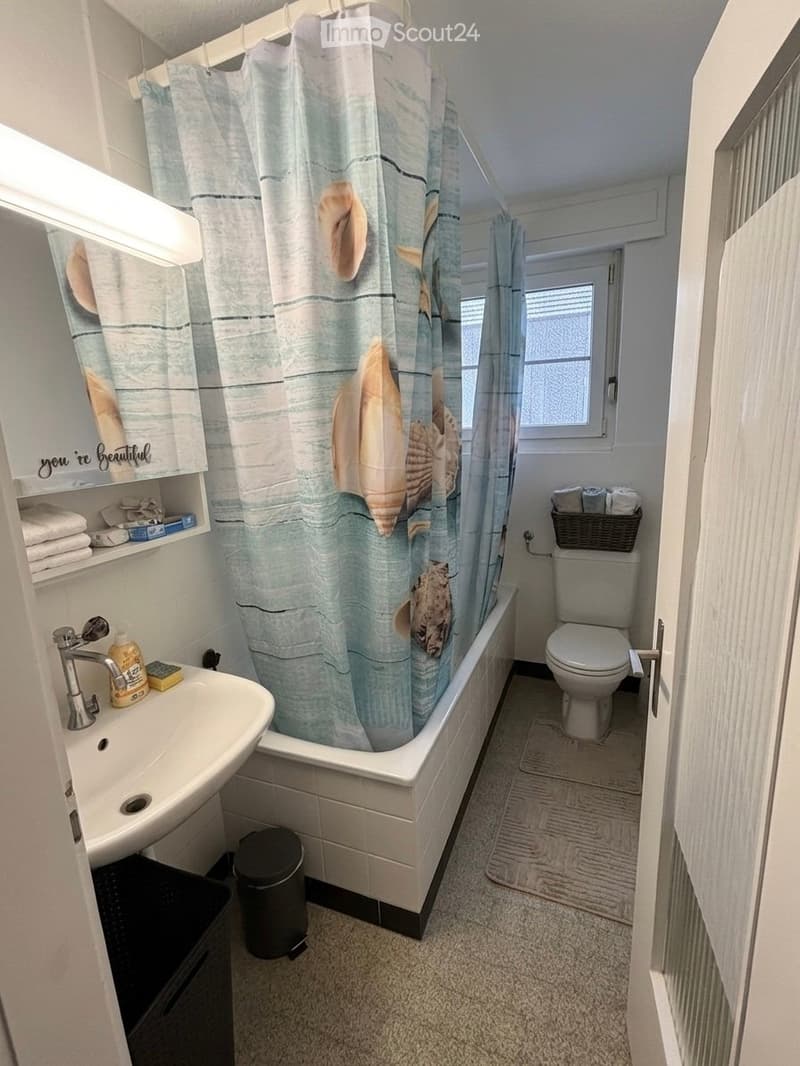 Cozy bathroom with a bathtub, toilet, sink, and shower curtain featuring seashells. Includes a towel rack, wicker basket, and a small trash can.