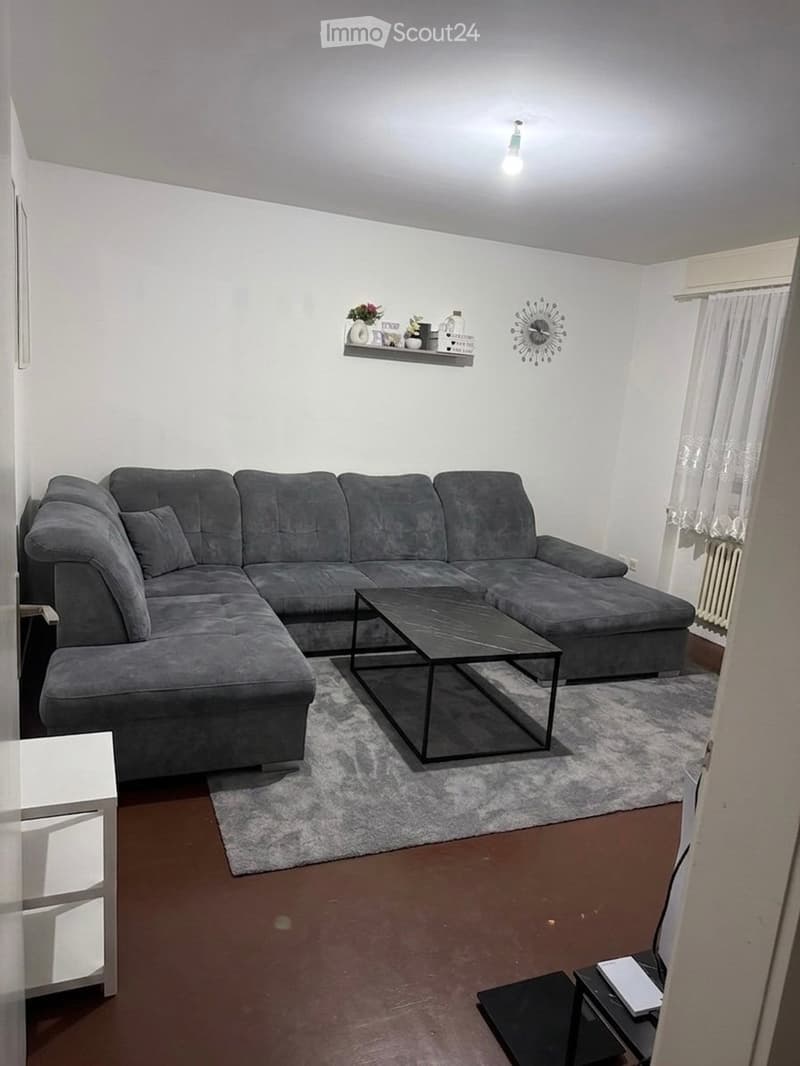 Modern living room with a grey sectional sofa, black coffee table, white shelf with decorative items, wall clock, side table, and curtained window.