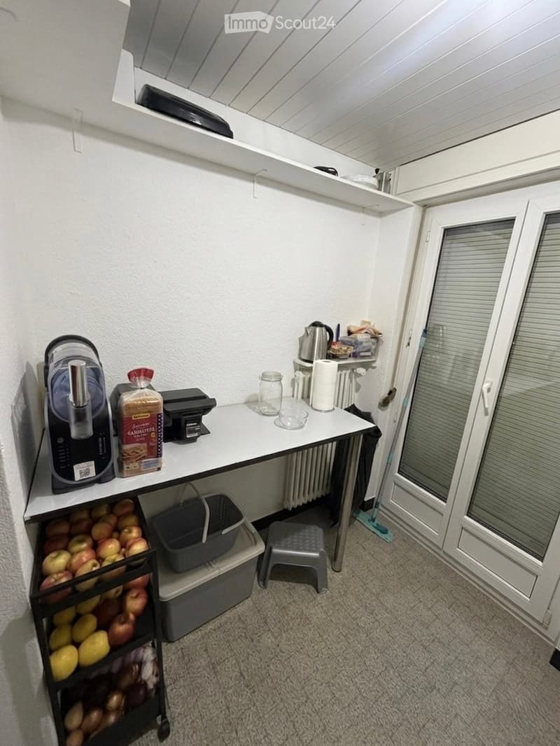 A small, well-organized kitchen area with a white table, a stool, a basket, a refrigerator, and various kitchen items including a cup, a bottle, and a small cabinet.