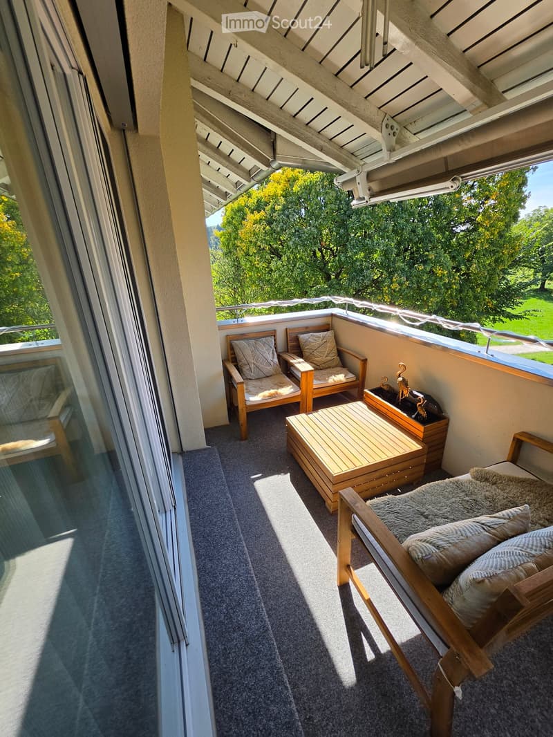 Balcony with wooden furniture including chairs, sofa, ottoman, and a small table, with a view of greenery outside.