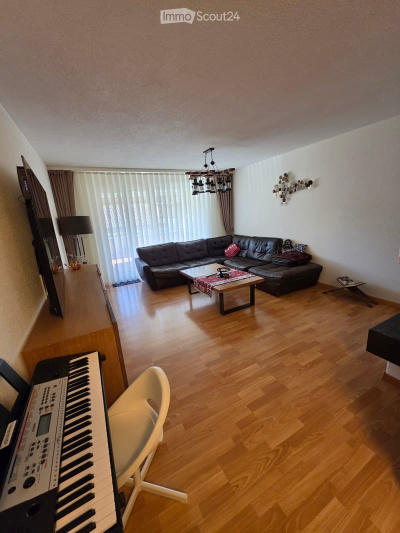 A modern living room with a black leather sofa, coffee table, piano, and a large window with vertical blinds.