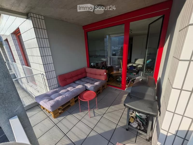 Balcony with a modern design, featuring a grey sectional sofa, a small pink table, and a grey barbecue. The space includes a tiled floor and a view of the city.