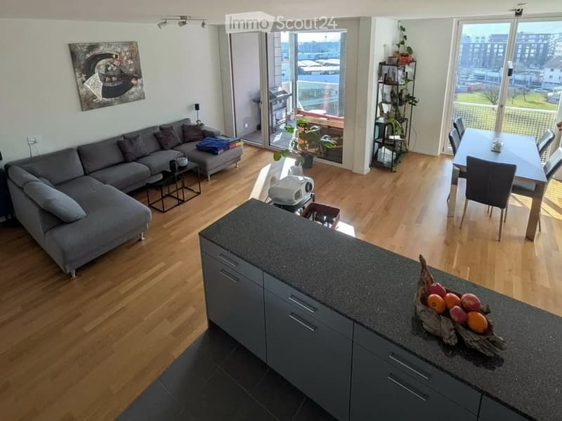 Modern, open-plan living and dining area with wooden flooring, grey sectional sofa, coffee table, dining table with chairs, kitchen island, and large windows offering a view of the city.