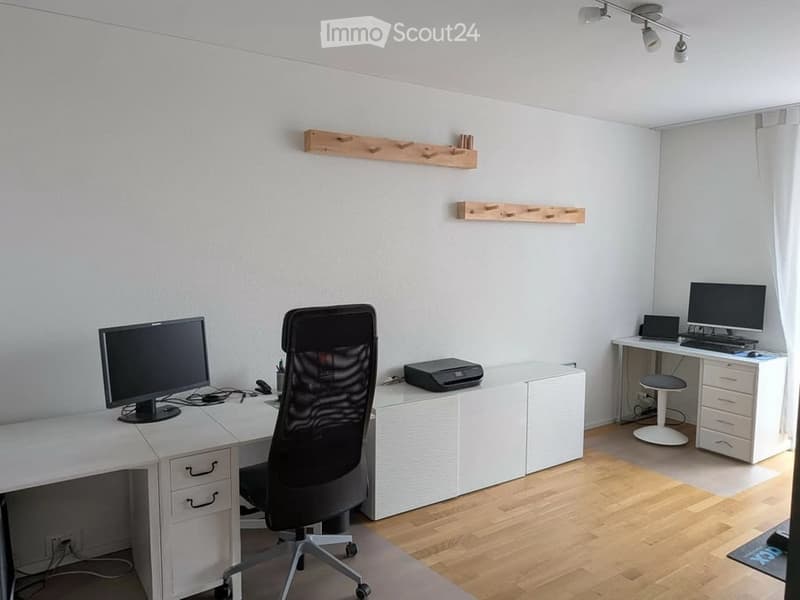 A modern, well-lit office space with white walls, wooden flooring, and multiple desks equipped with computer monitors, keyboards, mice, and a printer. A black office chair is positioned in front of one desk, and a small white stool is near another desk. W