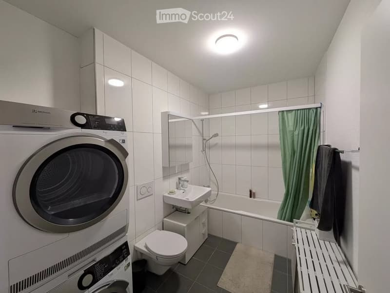 Modern bathroom with white tiled walls, a sink, toilet, bathtub with shower, mirror, washing machine, dryer, and a green shower curtain.