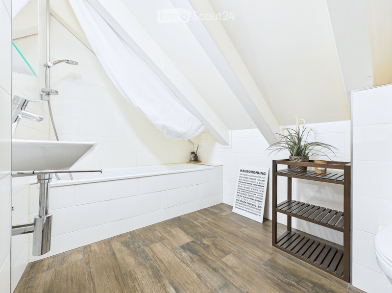 Modern bathroom with white tiled walls, wooden flooring, a bathtub, a sink with a modern faucet, a small wooden shelf with a plant, and a toilet.