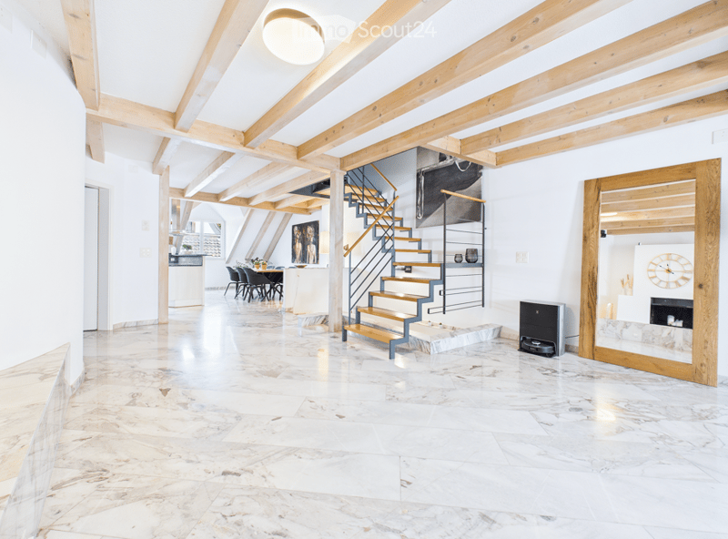 Bright, open hallway with exposed wooden beams, marble flooring, and a modern staircase leading to an upper level. The space features a dining area with chairs and a table visible in the background.
