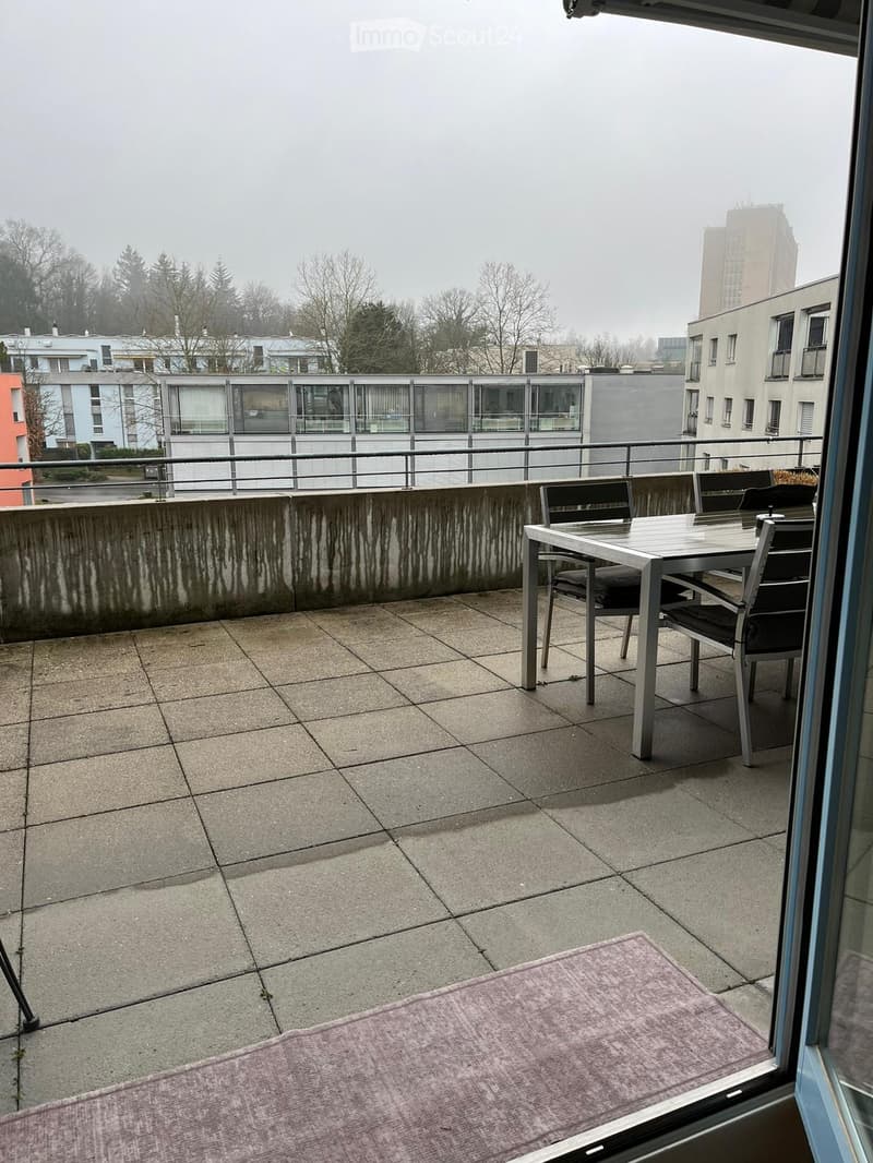 Balcony with tiled floor, metal table and chairs, and a view of surrounding buildings and trees.