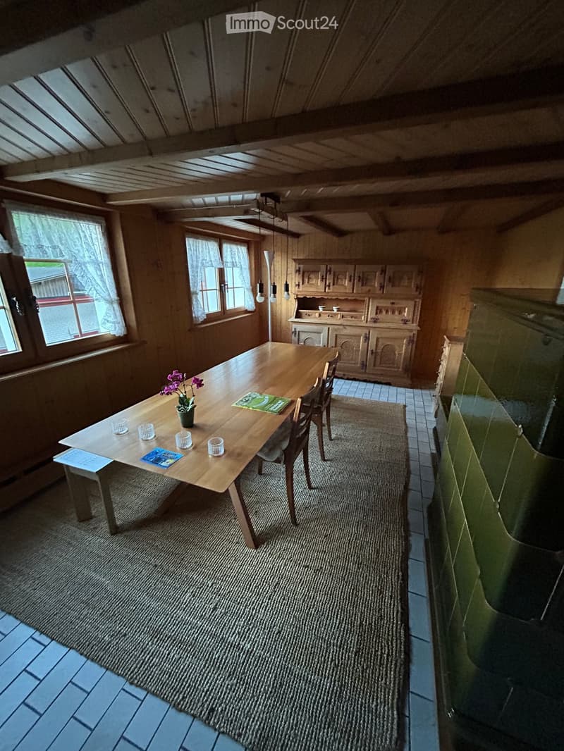 Cozy kitchen with wooden dining table, chairs, and a cabinet. The room features wooden walls, a tiled floor, and a large window with curtains.