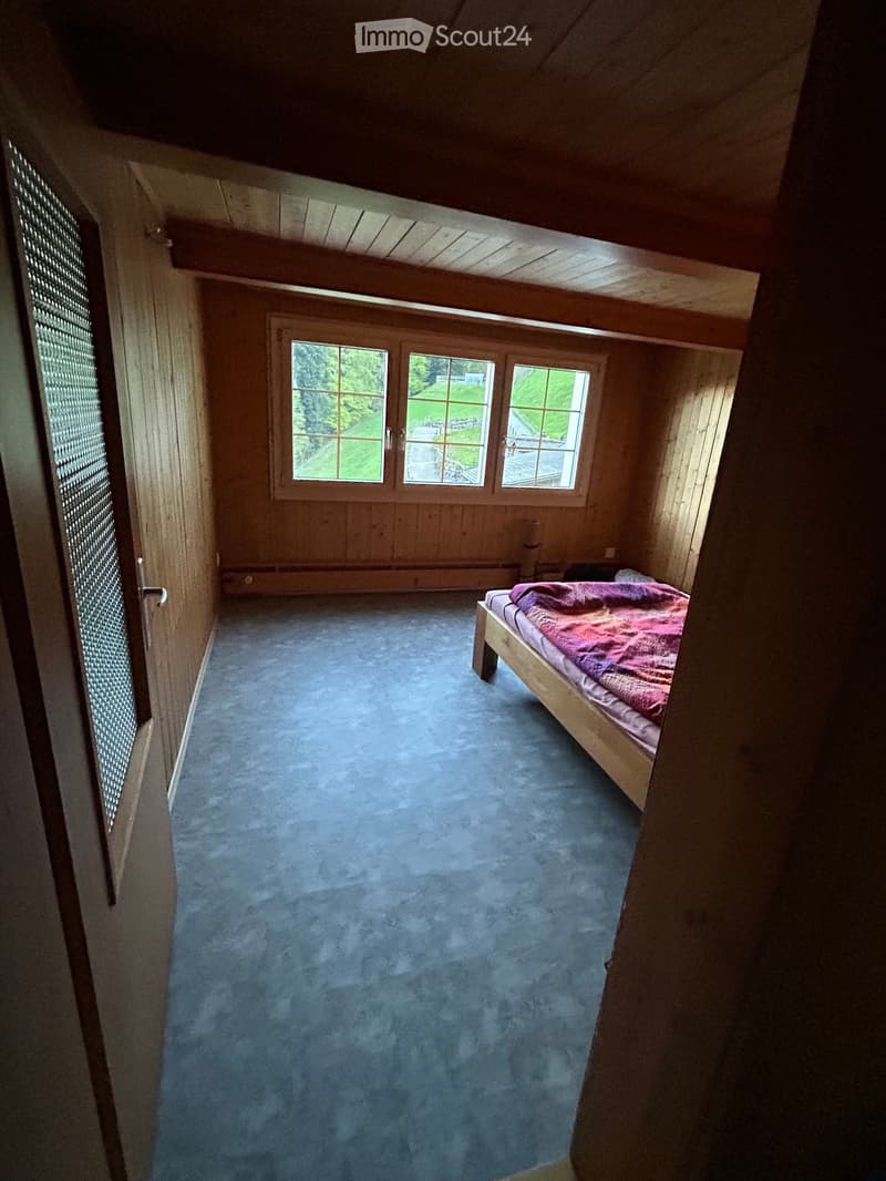 Cozy attic bedroom with wooden walls, slanted ceiling, blue carpet, and a single bed with a pink blanket.