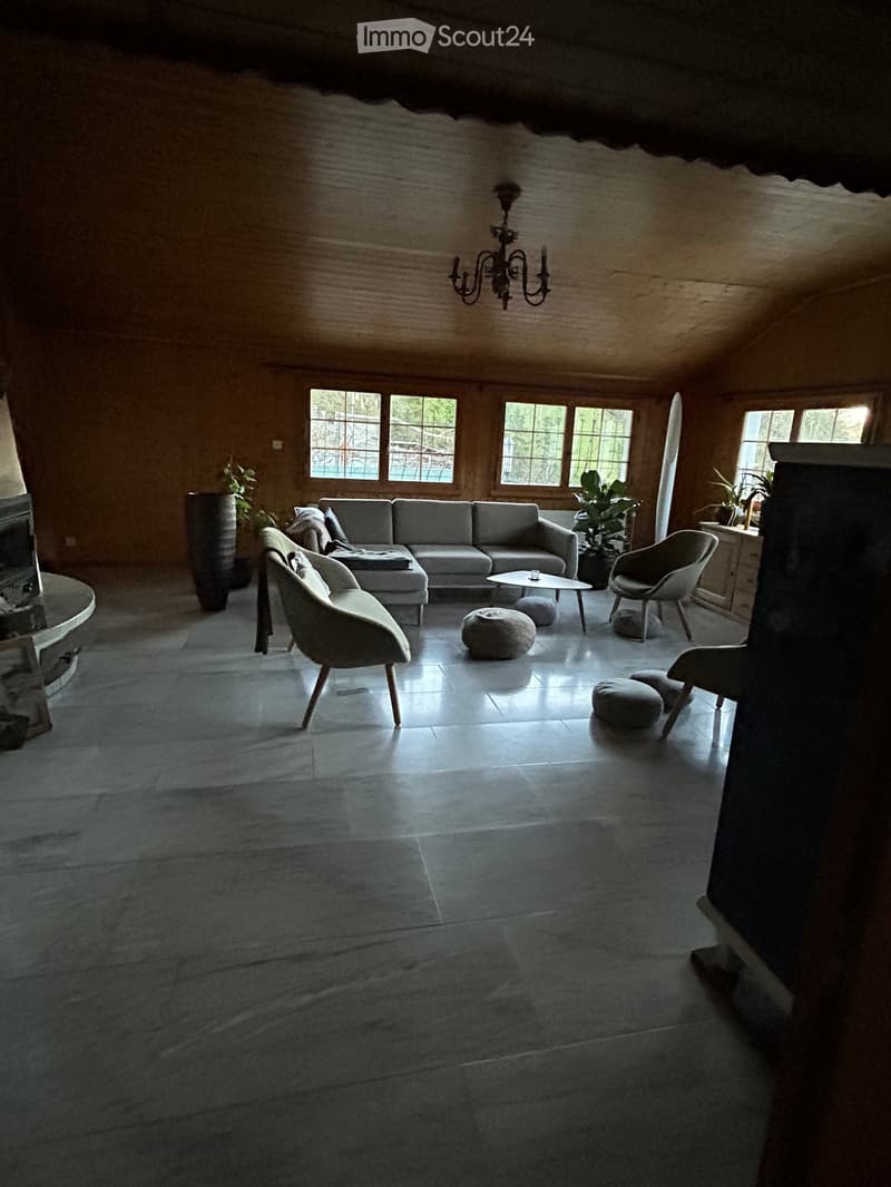 Spacious living room with wooden walls, large windows, a grey sectional sofa, coffee table, chairs, and a chandelier.