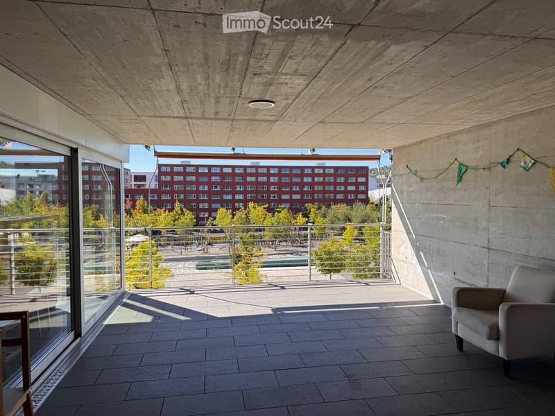 Modern balcony with concrete floors and walls, large glass windows, metal railing, and a view of a cityscape with trees and buildings.