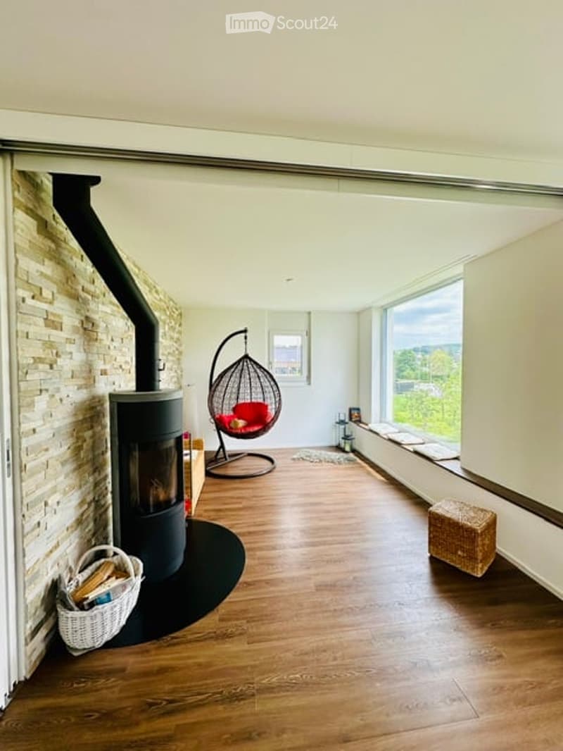 Modern living room with wooden flooring, large windows, a stone accent wall, a black fireplace, a red hanging egg chair, and a wicker basket with logs.