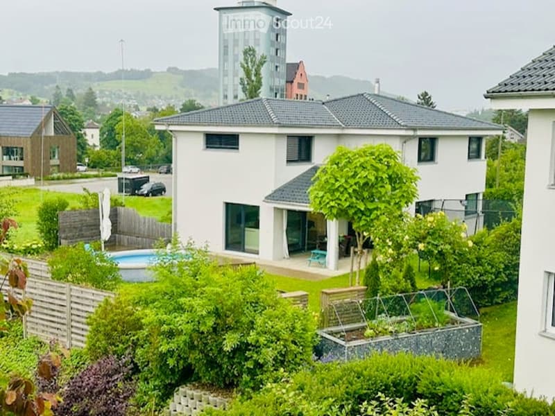 Modern two-story house with a grey roof, white walls, and large windows. Features a swimming pool in the garden, surrounded by greenery and a wooden fence.