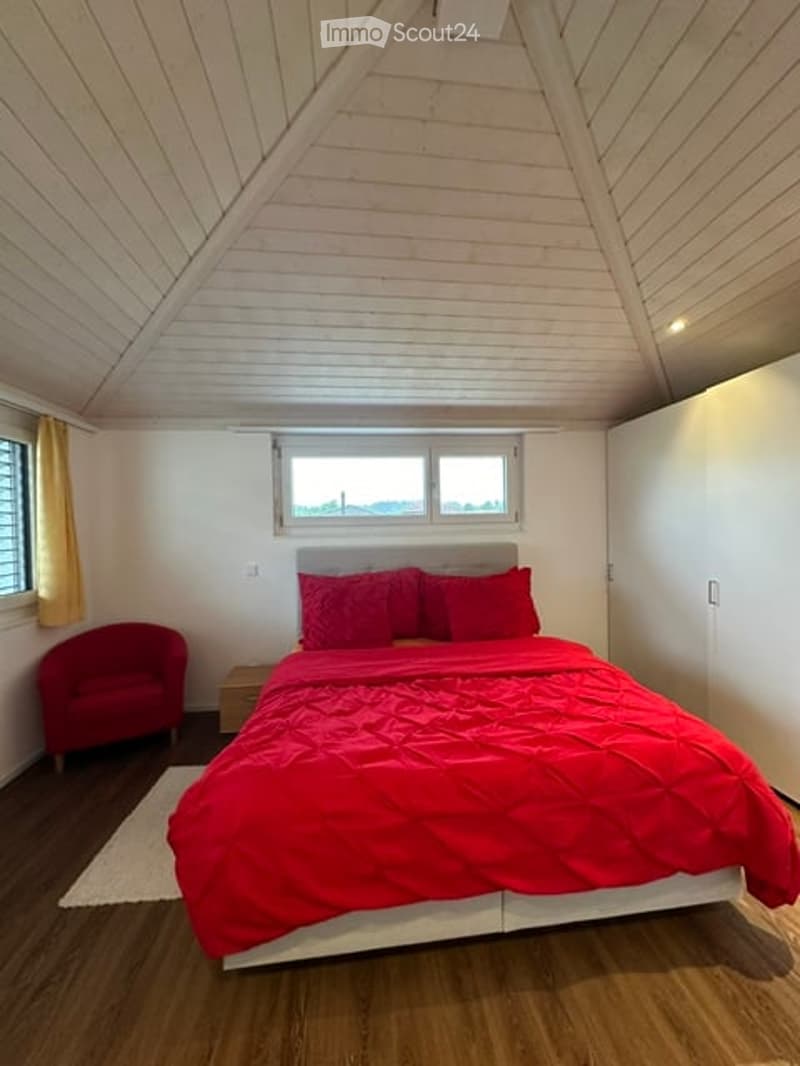 Cozy bedroom with a slanted wooden ceiling, large windows, red bedding, a red armchair, and a nightstand.