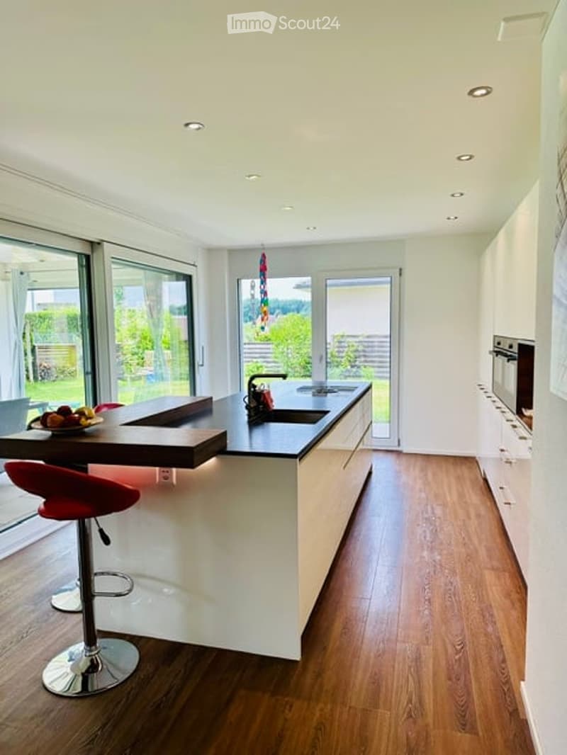Modern kitchen with white cabinets, dark countertops, island, red stools, and large windows overlooking a garden.