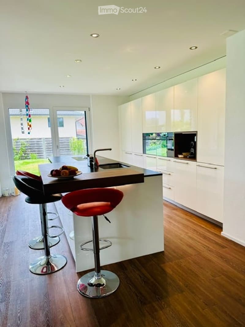 Modern kitchen with white cabinets, a central island, and red bar stools. Features a sink, oven, and a plate with fruit on the island.