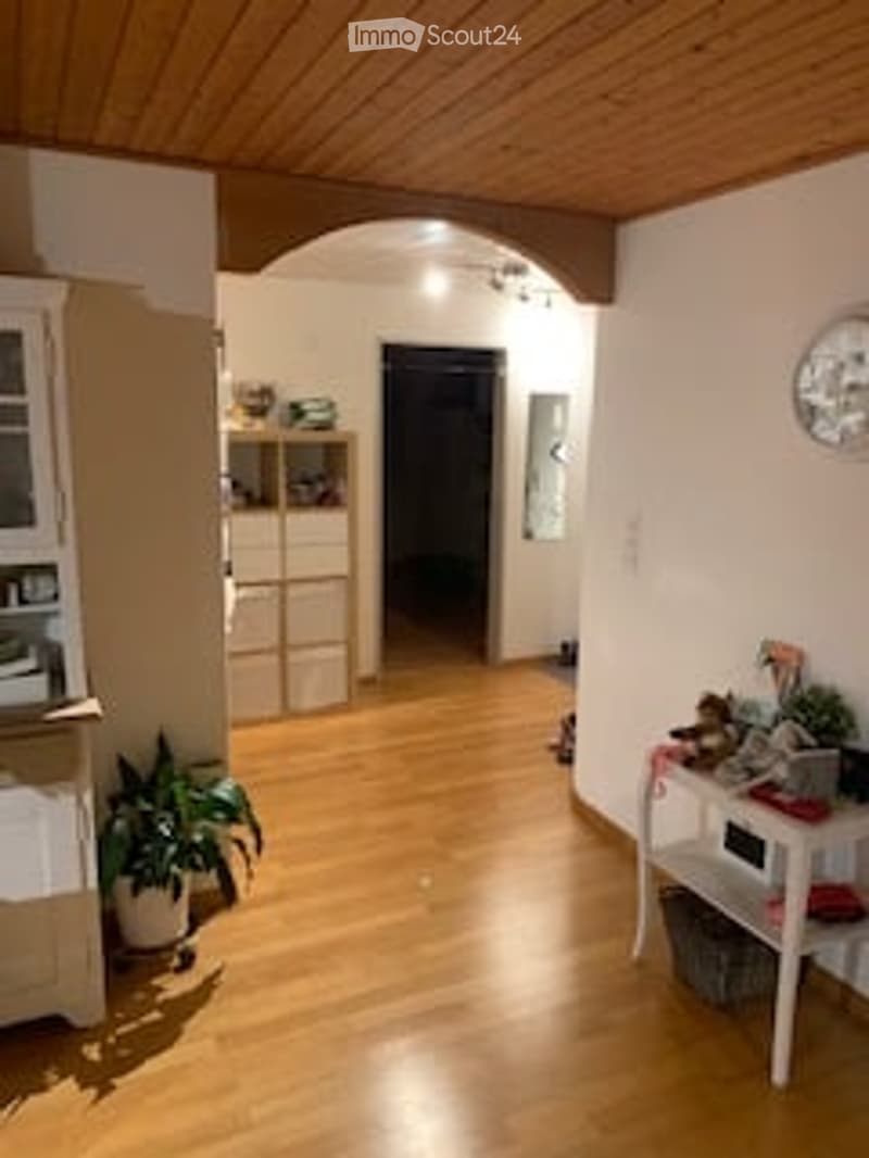 Bright hallway with wooden flooring, white walls, and a small table with plants. A cabinet and shelves are visible on the left side.