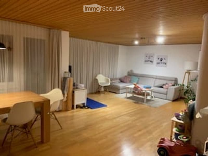 Bright, open-plan living and dining area with wooden flooring, large windows with curtains, grey sofa, coffee table, dining table with chairs, and decorative items.