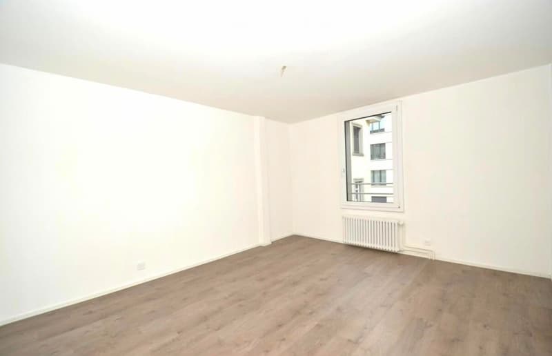 Empty room with light wooden flooring, white walls, and a single window with a radiator below it.