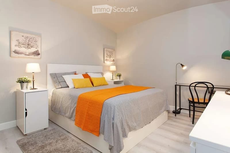 Modern bedroom with a large bed, nightstand, desk, and chair. Features a colorful bedspread, lamps, and artwork on the walls.