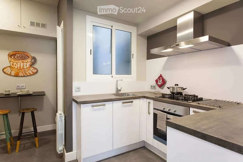 Modern kitchen with white cabinets, stainless steel appliances, a sink, stove, and oven. Includes a small table with two stools and a decorative wall sign.