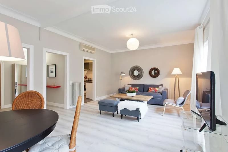 Modern living room with light grey walls, white flooring, and large windows. Features a blue sofa, wooden coffee table, ottoman, wicker chair, glass TV stand, and decorative mirrors.